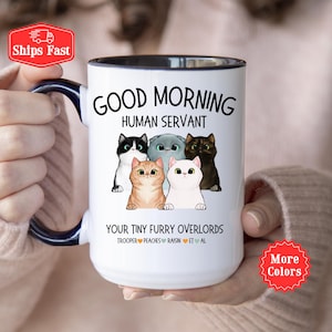 Puede incluir: Taza de caf&eacute; de cer&aacute;mica blanca con borde y asa negros. La taza presenta una ilustraci&oacute;n de dibujos animados de cinco gatos con el texto "Good Morning Human Servant" y "Your Tiny Furry Overlords Trooper Peaches Raisin Et Al".