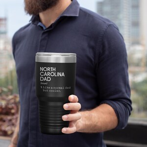 May include: Black insulated tumbler with a silver rim, featuring the text "NORTH CAROLINA DAD (noun) 1. Like a normal dad but cooler." The tumbler is held by a person. The tumbler is designed to keep beverages cold or hot.
