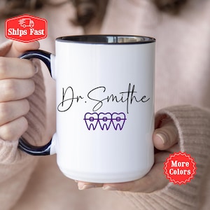 Puede incluir: Taza de cerámica blanca con borde y asa negros. La taza tiene un diseño morado de tres dientes con brackets y el texto "Dr. Smithe" en cursiva negra.