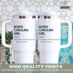 May include: Two white insulated tumblers with handles and lids. Each tumbler features the text "NORTH CAROLINA GIRL" with a definition. The tumblers have a silver rim and a white handle. A straw is visible in one of the tumblers.