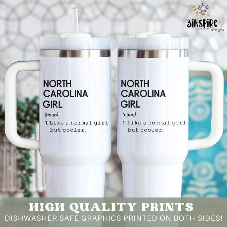 May include: Two white insulated tumblers with handles and lids. Each tumbler features the text "NORTH CAROLINA GIRL" with a definition. The tumblers have a silver rim and a white handle. A straw is visible in one of the tumblers.