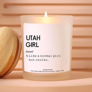 May include: A lit candle in a frosted glass jar with a label that reads "UTAH GIRL". The label also states "Like a normal girl but cooler." The candle is hand-poured soy wax and is 8 oz.