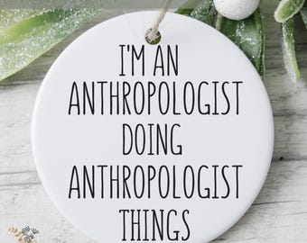 May include: A round white ceramic ornament featuring the black text I'M AN ANTHROPOLOGIST DOING ANTHROPOLOGIST THINGS. The ornament has a small hole at the top for hanging and is presented with a simple string loop for display purposes.