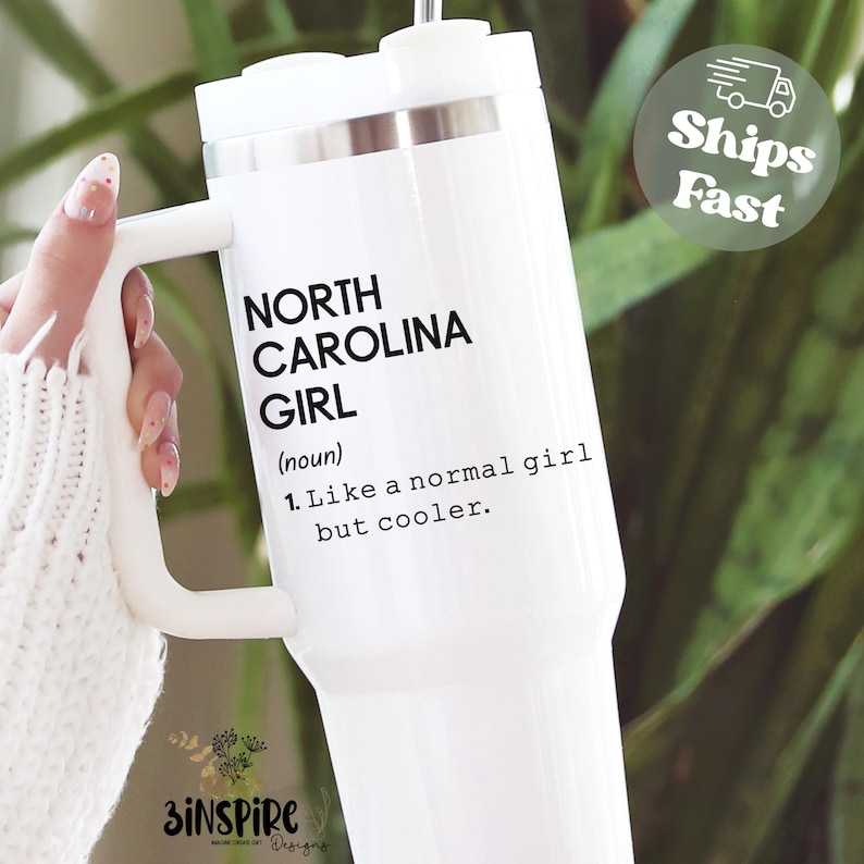 May include: White travel mug with a handle, featuring the text "NORTH CAROLINA GIRL" and a definition. The mug has a white lid and a silver band. The text reads "Like a normal girl but cooler."