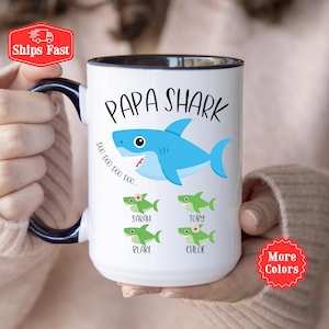 May include: White ceramic mug with a black interior and handle. The mug features a blue shark illustration with the words "PAPA SHARK" and the names "Sarah", "Toby", "Blake", and "Chloe" below. The mug also has the phrase "Doo Doo Doo Doo..." printed on it.