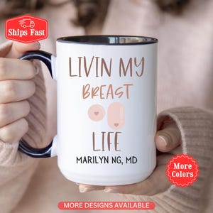 Könnte beinhalten: Ein weißer Keramikbecher mit dunkelblauem Henkel und Rand. Der Becher zeigt den braunen Schriftzug "LIVIN MY BREAST LIFE" über zwei hellrosa kreisförmigen und ovalen Formen mit kleinen Herzen in der Mitte sowie den schwarzen Text "MARILYN NG, MD".