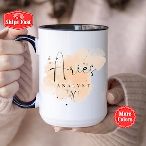 May include: A white ceramic mug with a dark blue handle and interior rim. The front features a peach-colored watercolor splash design with the text Aries, the word ANALYST in capital letters, and the Aries zodiac symbol printed in black ink.