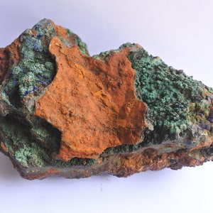 Puede incluir: Formación rocosa natural con una mezcla de colores. Los tonos dominantes son el naranja óxido y el verde intenso, con toques de azul y marrón. La textura parece áspera y desigual, sugiriendo un espécimen mineral natural.