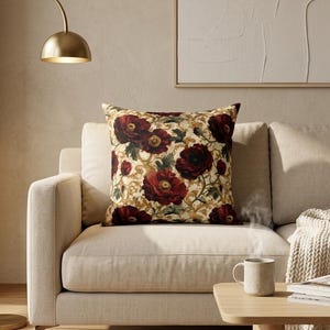 Op de afbeelding: Een vierkant sierkussen met een herhalend patroon van donkerrode klaprozen en donkergroen blad op een crèmekleurige achtergrond, voorzien van gedetailleerde goudkleurige krulmotieven, geplaatst op een bank in neutrale tinten.