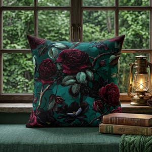 May include: A square throw pillow featuring a dark teal background with a dense pattern of deep red roses and green leaves. The pillow is positioned next to two stacked vintage books and a brass-colored metal lantern with a glowing light.