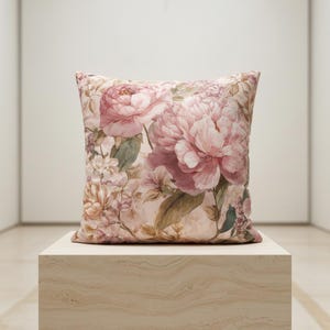 May include: Square decorative pillow featuring a large floral pattern of pink peonies and other flowers on a light pink background. The pillow is placed on a light-colored, textured surface.