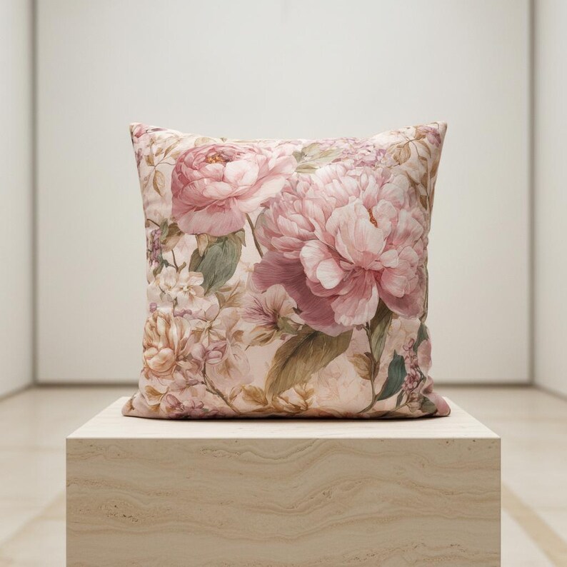 May include: Square decorative pillow featuring a large floral pattern of pink peonies and other flowers on a light pink background. The pillow is placed on a light-colored, textured surface.