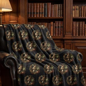 May include: A black throw blanket draped over a leather armchair. The blanket features a repeating pattern of a rabbit surrounded by a wreath of flowers and foliage. The background includes a wooden bookshelf and a lamp.