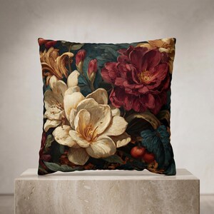 May include: A square throw pillow featuring a dark, moody floral print. The design displays large, cream-colored lilies, a deep burgundy peony, and smaller red buds set against a dark teal and green foliage background with small red berries.