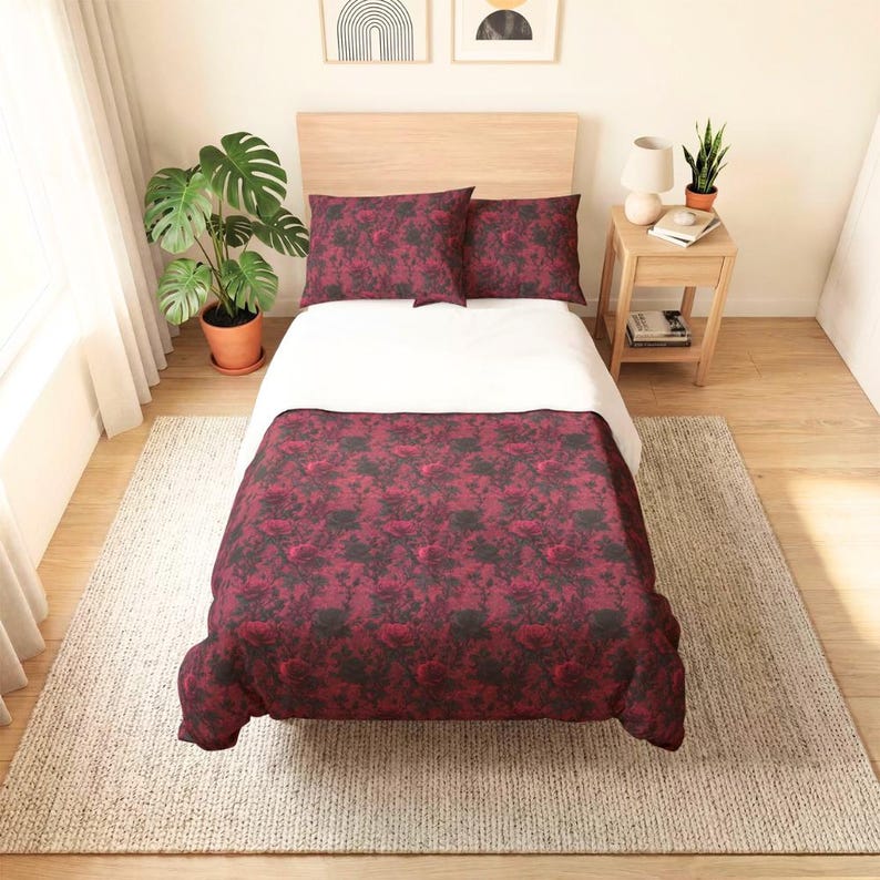Pu&ograve; includere: Un letto con un piumino e due cuscini con un motivo floreale rosso scuro e nero. Il letto presenta un lenzuolo bianco, una testiera in legno chiaro ed &egrave; posizionato su un tappeto di colore chiaro in una stanza con una pianta e un comodino.