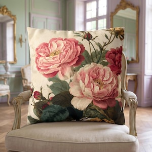 May include: A square throw cushion featuring a botanical illustration of large, blooming pink and red peonies with dark green leaves and small buds on a cream-coloured background. The floral design covers the front of the cushion.