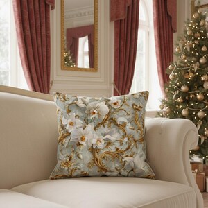 May include: A square throw pillow featuring a light blue background with a repeating pattern of white orchid flowers and intricate gold scrolling filigree designs. The pillow has a soft fabric texture and is displayed on a neutral-colored sofa.