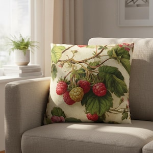 May include: Square decorative pillow with a botanical raspberry design. The pillow features a cream-colored background with a detailed illustration of red and yellow raspberries, green leaves, and delicate branches. The pillow is placed on a beige sofa.