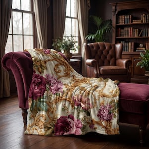 Op de afbeelding: Een decoratieve deken met bloemenpatroon in bordeaux, roze en groen met gouden versieringen, gedrapeerd over een bordeaux fluwelen chaise longue. Het bloemmotief is in barokstijl. De kamer heeft een houten vloer.