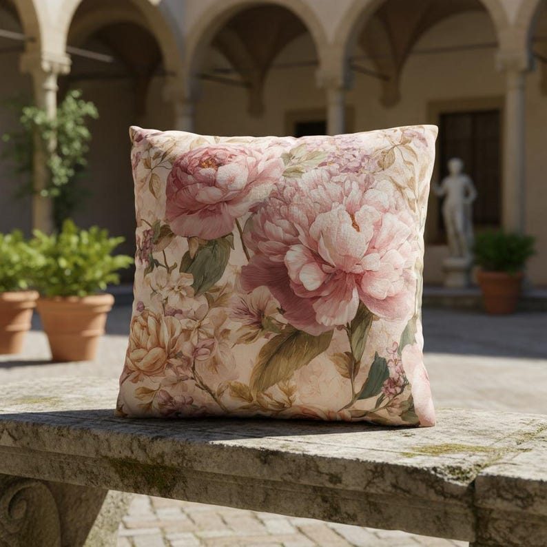 May include: Square decorative pillow featuring a large floral pattern of pink peonies and other flowers on a cream background. The pillow is resting on a stone surface, suggesting outdoor use.