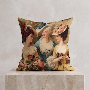 May include: Decorative square pillow featuring a vintage painting of three women in elegant attire. The women are wearing hats adorned with flowers and lace-trimmed dresses in shades of blue, yellow, and cream. The pillow is placed on a marble surface.