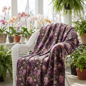 May include: A white wicker chair draped with a plush, floral-patterned blanket. The blanket features a deep purple background with pink and peach rose blooms and green foliage. The scene is set in a sunroom with potted orchids and ferns.