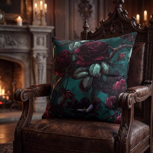 May include: Square decorative pillow with a dark teal background and a floral pattern of large, deep red roses and green leaves. The pillow is placed on a brown leather armchair with a carved wooden frame.