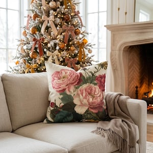 May include: A square throw pillow featuring a botanical print of large, layered pink and deep red peonies with dark green foliage on a cream-colored background, resting on a light beige sofa with a matching throw blanket.