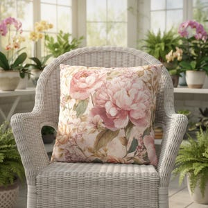 May include: A square decorative pillow with a floral pattern featuring large pink peonies and smaller white flowers. The pillow is set on a white wicker chair in a sunroom with potted plants.