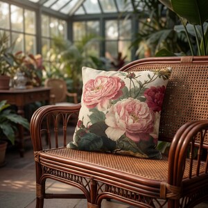 May include: A square scatter cushion featuring a botanical print of large, layered pink and deep red peonies with green leaves on a cream-coloured background, resting on a brown woven rattan bench with a curved back and armrests.