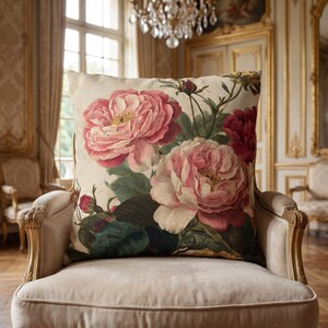 May include: A square throw pillow featuring a botanical illustration of large, blooming pink peonies with dark green leaves and small red buds on a cream-coloured background. The floral design covers the front surface of the pillow.