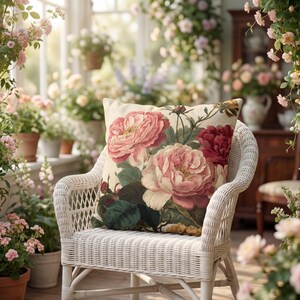 May include: A square throw pillow featuring a botanical print of large pink and deep red roses with green leaves on a cream-coloured background. The pillow is placed on a white wicker chair, which is positioned in a sunlit room filled with potted flowers.