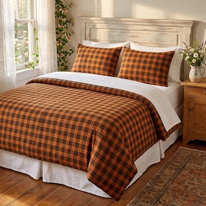 Puede incluir: Una cama con un edredón y dos fundas de almohada a juego con un patrón de cuadros naranja y marrón. La ropa de cama se coloca sobre un volante de cama blanco y sábanas blancas, frente a un cabecero de madera blanca en un dormitorio luminoso.