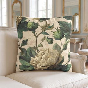 May include: Square throw pillow with a botanical design. The pillow features a large, detailed illustration of a peony flower and buds in shades of green and cream against a beige background. The pillow is placed on a white sofa.