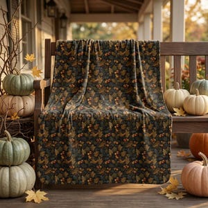 May include: A dark-colored throw blanket featuring a repeating pattern of brown acorns and golden-brown oak leaves. The blanket is draped over a wooden bench, surrounded by several pumpkins in shades of green, orange, and cream on a porch.