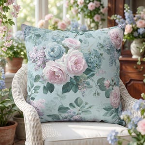May include: Square cushion with a floral pattern. It displays large pink and blue roses, lilac, and green foliage on a pale blue backdrop. The cushion is placed on a white wicker chair.