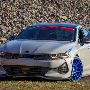 May include: A silver Kia K5 sedan with a black front bumper and blue and black wheels. The car has a red and black "KITZ" sticker on the windshield.