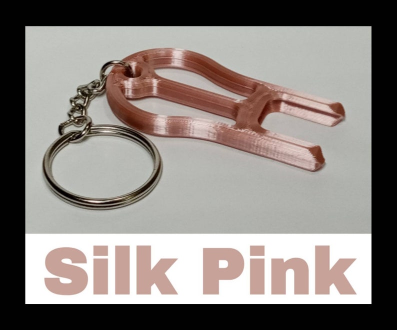 May include: A rose gold-colored keychain with a loop and a small, decorative, 3D printed object attached. The object is shaped like a stylized letter "H". The text "Silk Pink" is printed below the image.
