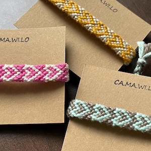 Puede incluir: Tres pulseras de la amistad en cartulina marrón con el texto "CA.MA.WILO" impreso en cada tarjeta. Las pulseras están tejidas con hilo amarillo y blanco, hilo rosa y blanco e hilo azul claro y gris.