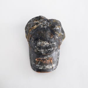 May include: A black baseball cap with a distressed, weathered finish. The cap has a curved peak and is covered in splatters of white, gold, and brown paint, creating a worn, vintage aesthetic. The background is plain white.