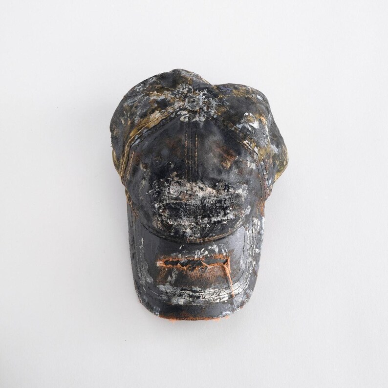 May include: A black baseball cap with a distressed, weathered finish. The cap has a curved peak and is covered in splatters of white, gold, and brown paint, creating a worn, vintage aesthetic. The background is plain white.