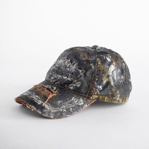 May include: A distressed baseball cap with a worn, textured appearance. The cap features a dark base colour with splattered white, silver, and brown paint. The brim has a frayed edge, adding to the vintage aesthetic.