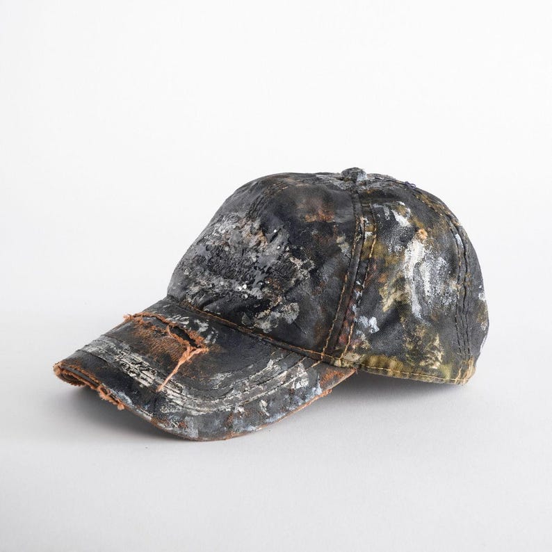 May include: A distressed baseball cap with a worn, textured appearance. The cap features a dark base colour with splattered white, silver, and brown paint. The brim has a frayed edge, adding to the vintage aesthetic.