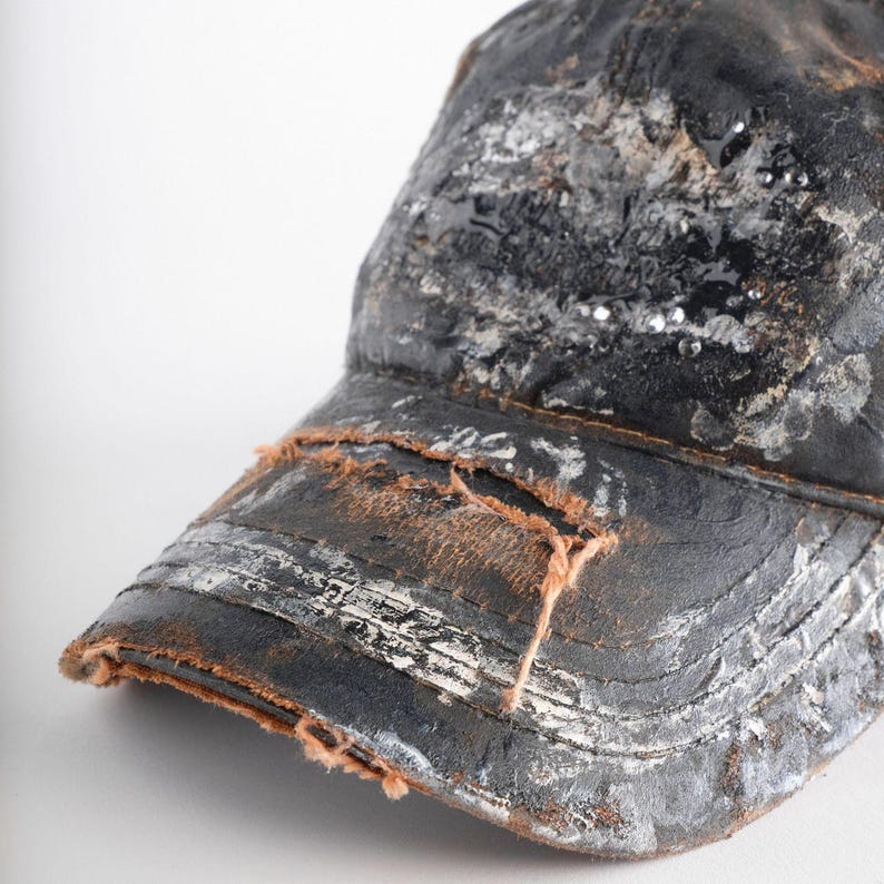 May include: A distressed black baseball cap with white and silver paint splatters. The brim has a worn, frayed edge. Small, shiny embellishments are scattered across the cap's surface. A classic, casual accessory.