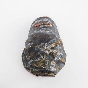 May include: A distressed black baseball cap with white and olive-green paint splatters. The cap has a brown leather brim and a small tear near the top. The overall look is worn and textured.