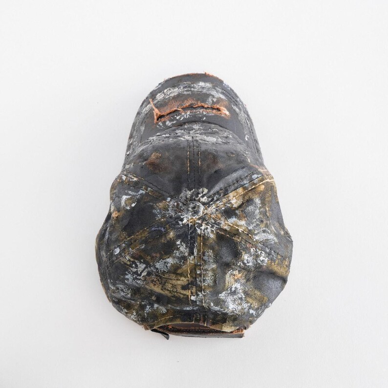 May include: A distressed black baseball cap with white and olive-green paint splatters. The cap has a brown leather brim and a small tear near the top. The overall look is worn and textured.