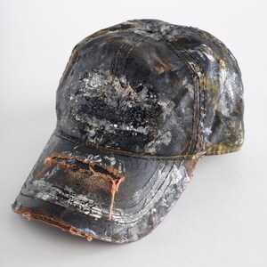 May include: A distressed black baseball cap with a curved peak. The cap has a worn, textured appearance with patches of white and silver paint. The peak has a distressed, torn edge.