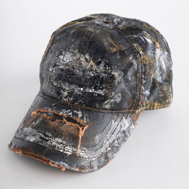 May include: A distressed black baseball cap with a curved peak. The cap has a worn, textured appearance with patches of white and silver paint. The peak has a distressed, torn edge.