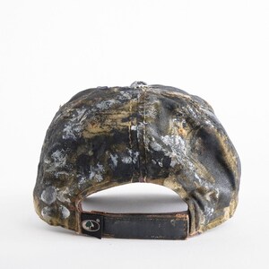 May include: A camouflage baseball cap with a black leather strap and a metal buckle. The cap features a pattern of black, brown, and white paint splatters. The back of the cap has a logo patch.