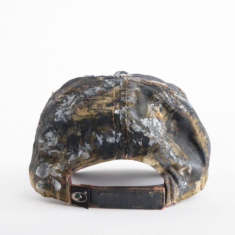 May include: A camouflage baseball cap with a black leather strap and a metal buckle. The cap features a pattern of black, brown, and white paint splatters. The back of the cap has a logo patch.